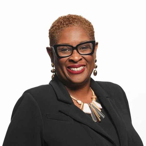 Smiling woman in black blazer with statement necklace and glasses.