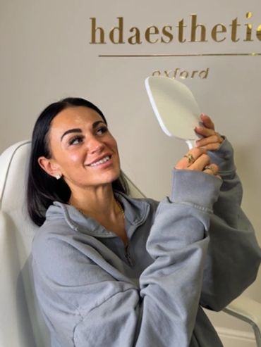 A woman in a gray hoodie smiles while looking in a handheld mirror.