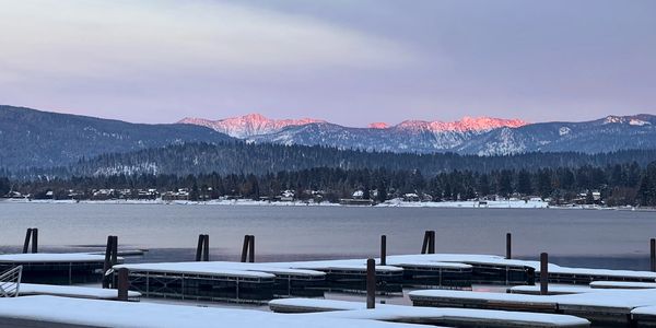 Snow-covered docks by a serene lake with pink-lit mountain peaks at dawn.