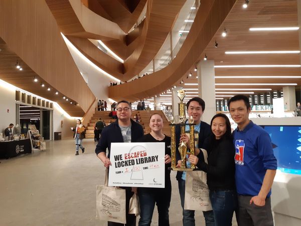 Group celebrates escape from locked library with sign and trophy in modern building.