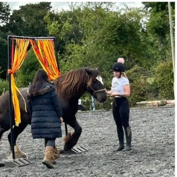Two women guiding a horse through a colorful curtain in an outdoor arena.