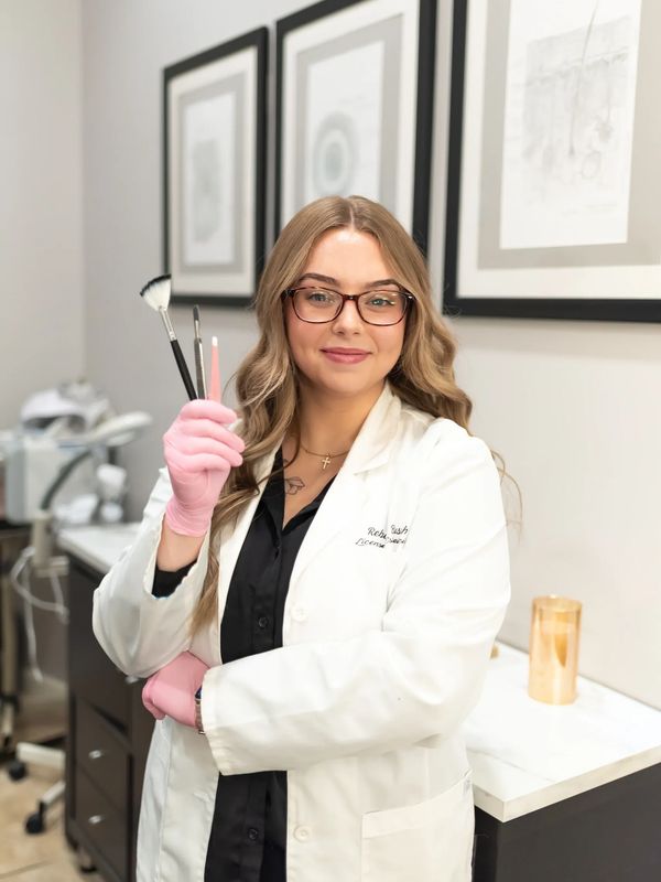 A smiling woman in a white lab coat holding makeup brushes in a clinical setting.