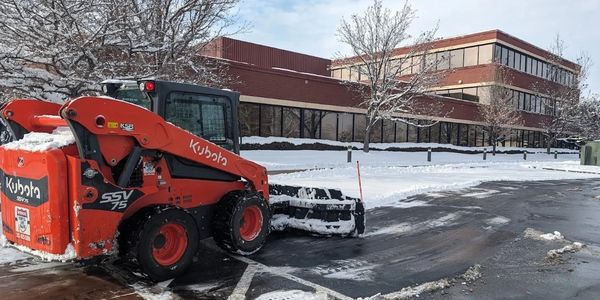 Kubota skid steer clearing snow in a parking lot near a brick building doing ice control, salting, salt, snow removal, 