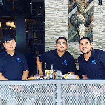 Three men smiling at a table in a restaurant, wearing matching navy shirts.
