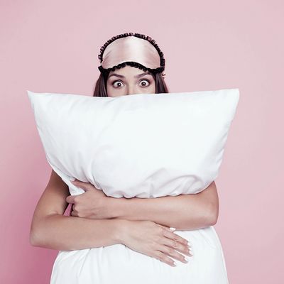 Woman with sleep mask hugging a large pillow, peeking with wide eyes.