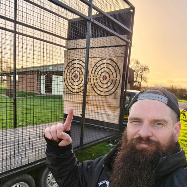 Man with a beard pointing at axe throwing targets in a fenced area.
