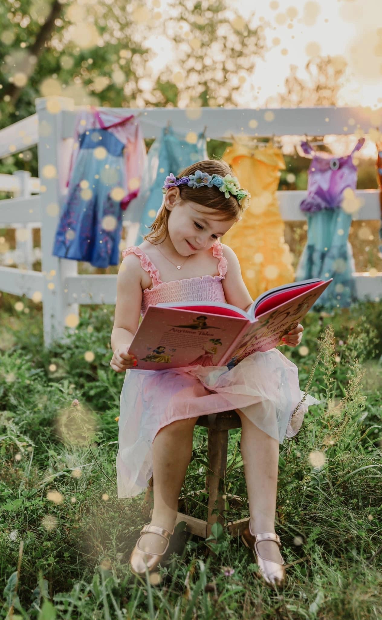 Young girl reading a book outdoors with dresses hanging behind her.