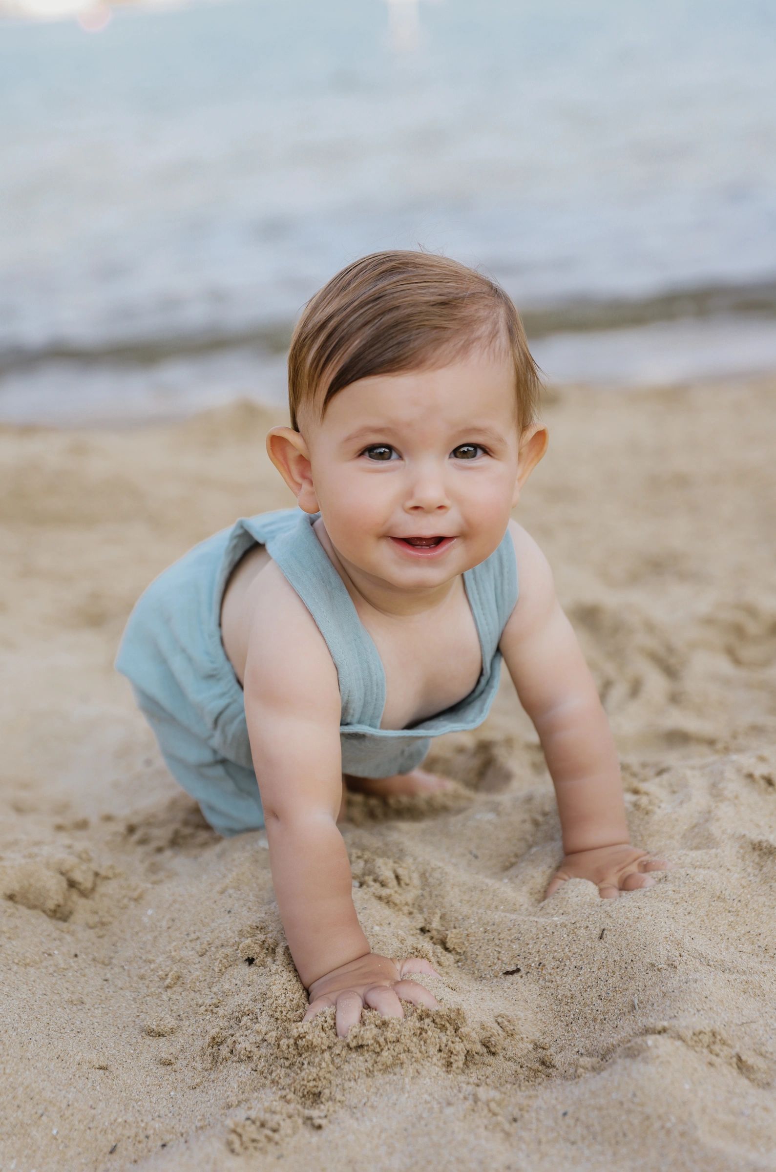 Smiling baby crawling on sandy beach near water.