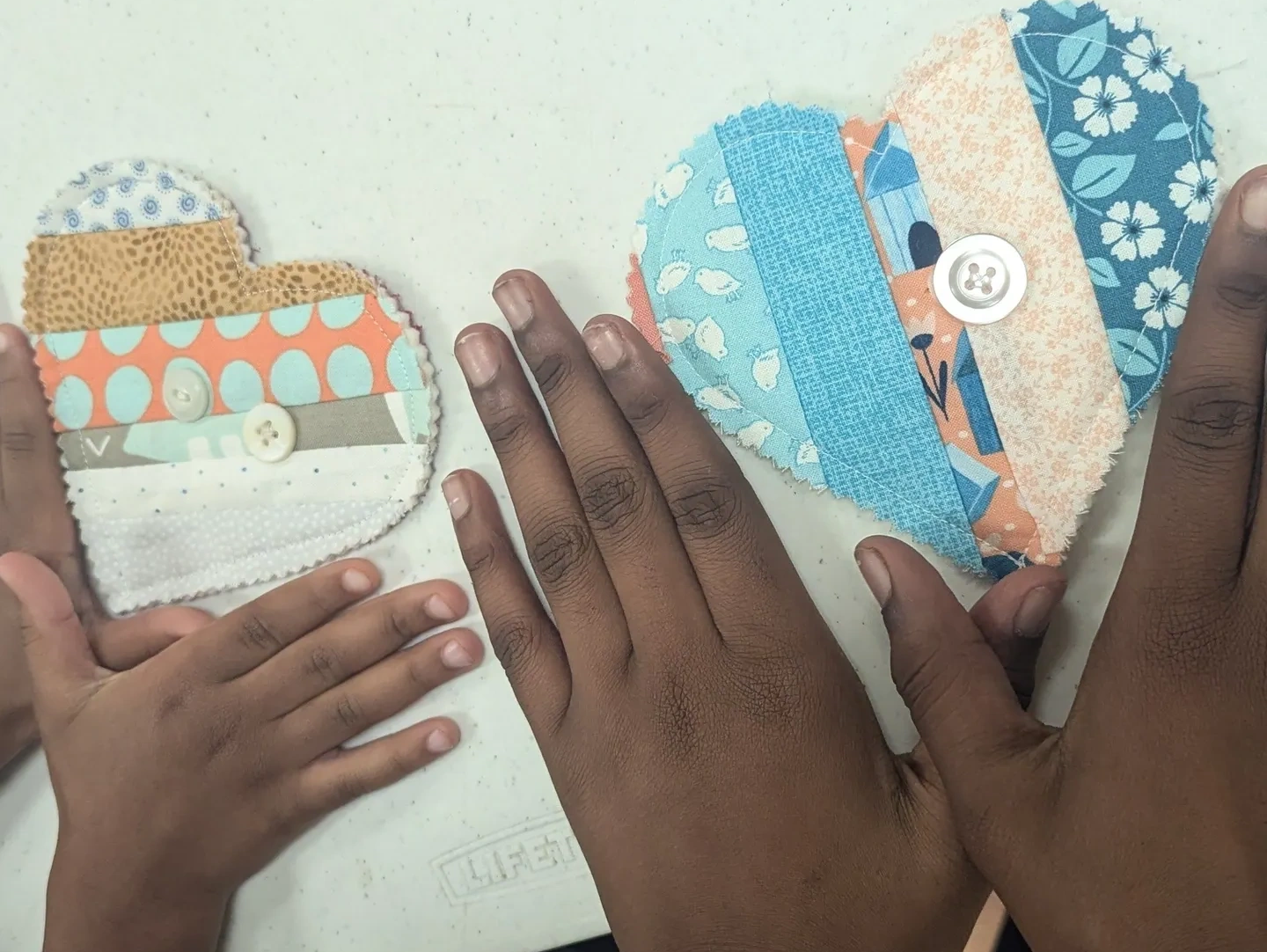 Two hands beside colorful patchwork heart-shaped fabric pieces with buttons.
