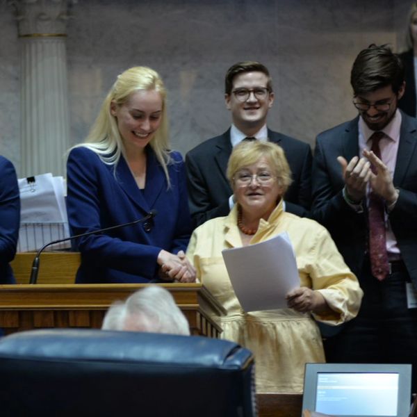 Two women shaking hands in a formal setting with people smiling.