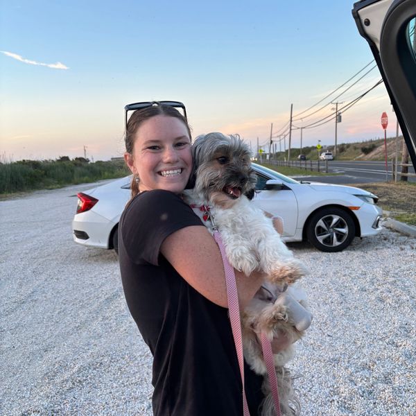 A smiling woman hugs her small dog near parked cars at sunset.