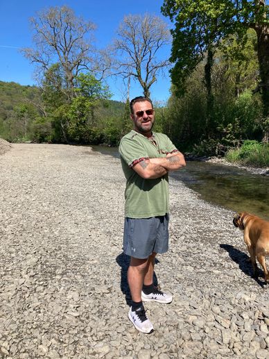 Man with sunglasses standing on rocky riverbank with dog nearby.