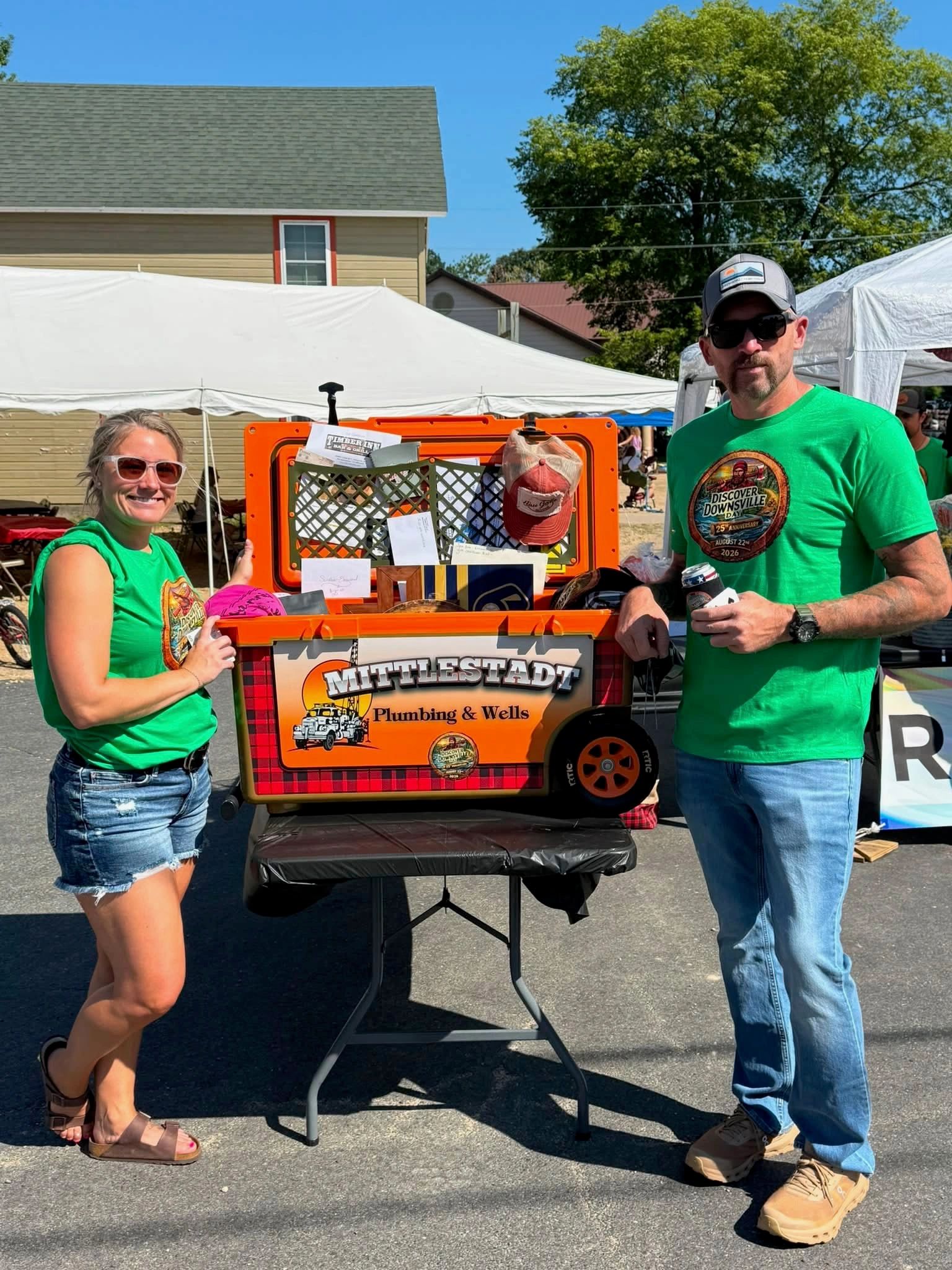 Two people stand beside a promotional cooler at an outdoor event.