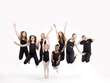 A group of seven girls jumping joyfully in dance attire against a white background.