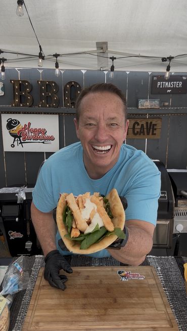 A smiling man presents a delicious taco with lettuce and sauce at a BBQ stall.