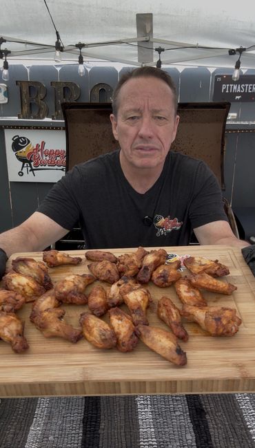 Man holding a board of cooked chicken wings at a BBQ setup.