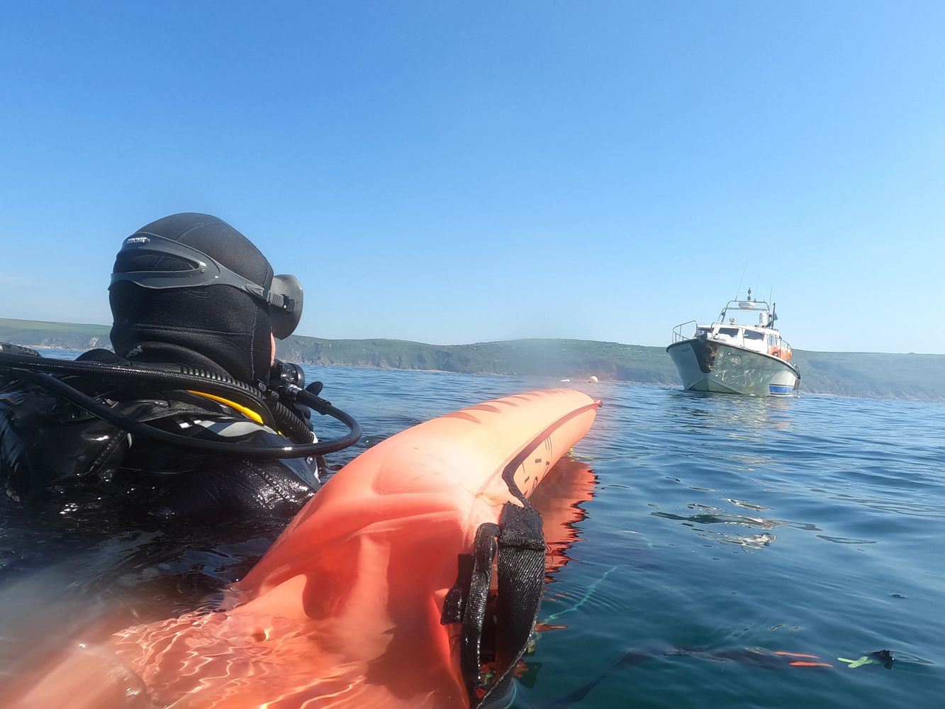 A diver in black gear floating near an orange buoy with a boat ahead on calm water.