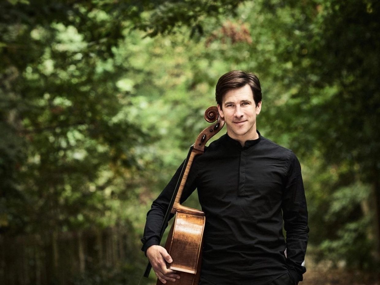 Man in black holding a cello in a forest path.