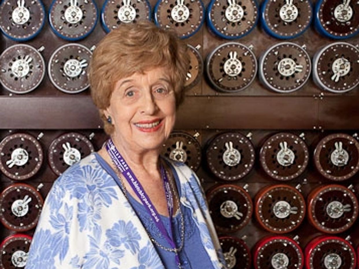 An elderly woman smiling in front of a vintage Enigma machine.