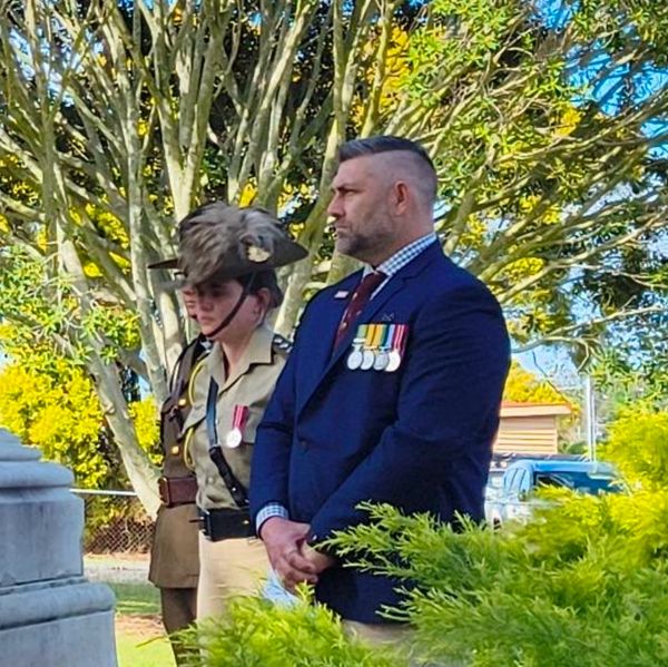 Two men in military and formal attire with medals stand solemnly outdoors near a monument.