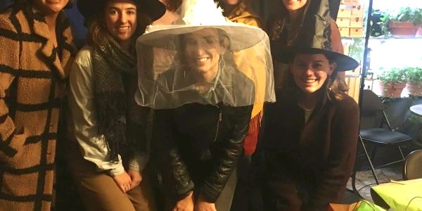 A group of women dressed in witch hats, the one front-and-center wearing a white "bride witch" hat w