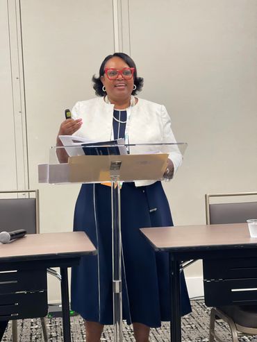 Woman in white jacket and red glasses giving a presentation at a podium.