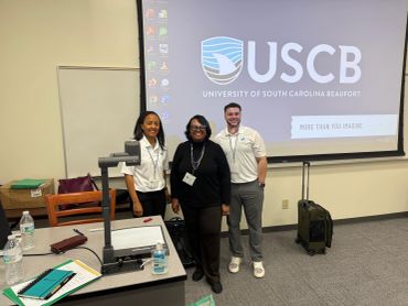 Three people posing in a classroom with a USC Beaufort presentation on screen.