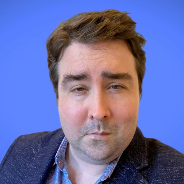 Man with brown hair wearing a blue blazer and patterned shirt against a blue background.