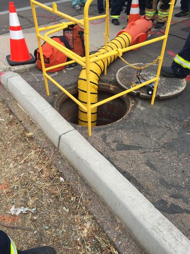 Safety barriers and ventilation hose set up around an open manhole on a street.