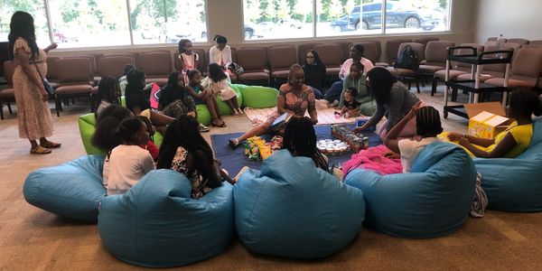 Children and adults gather indoors, sitting on bean bags and chairs during an activity session.