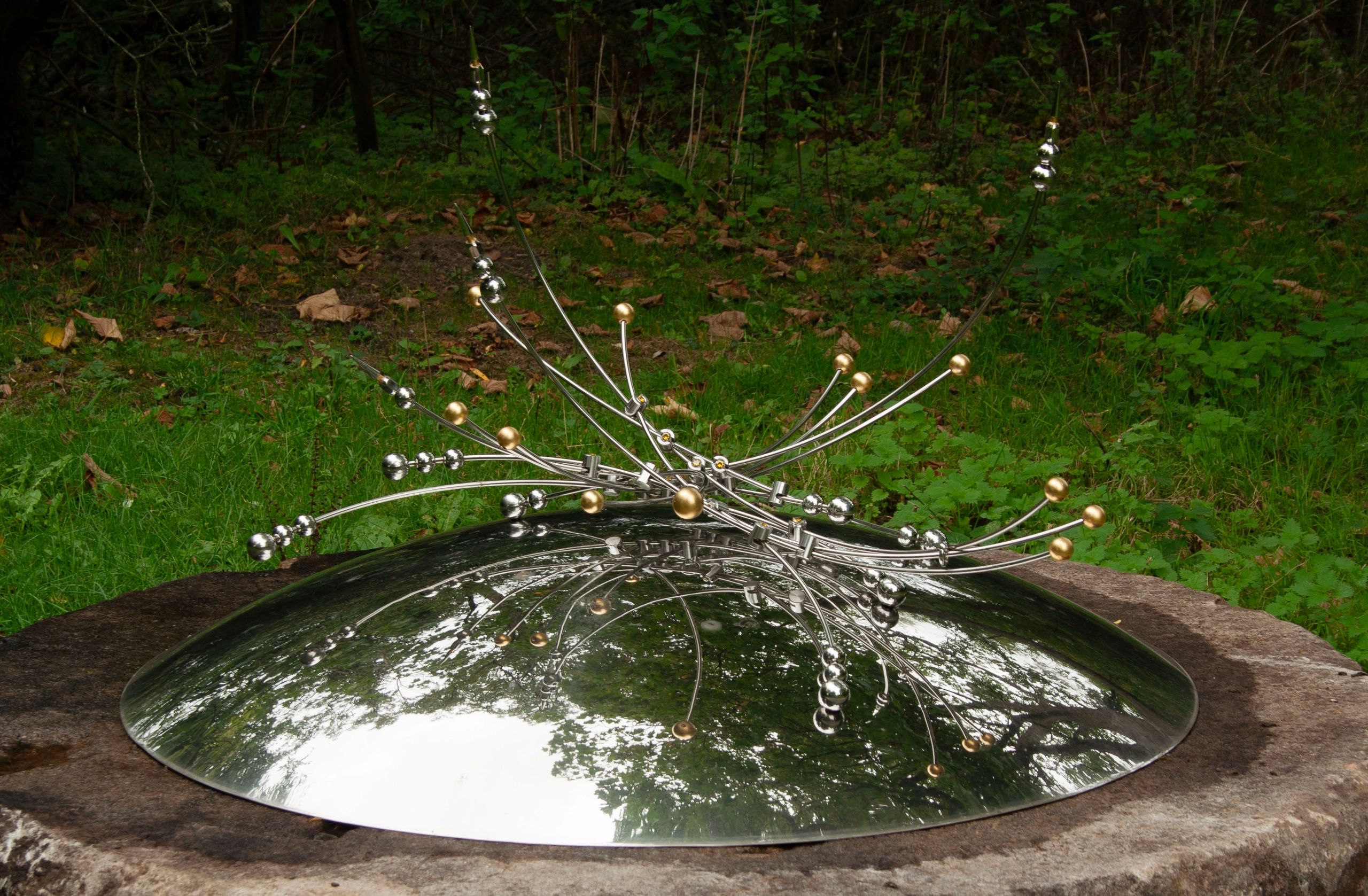Modern metallic sculpture with reflective dome and intricate rods in a natural setting.