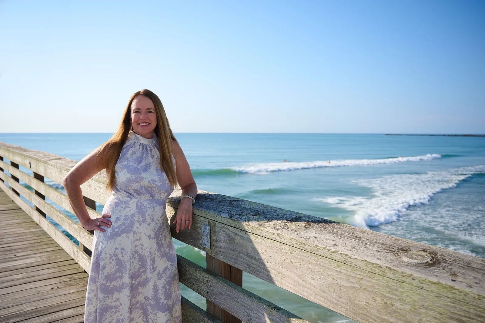 Woman in a floral dress smiling on a wooden pier by the ocean.
