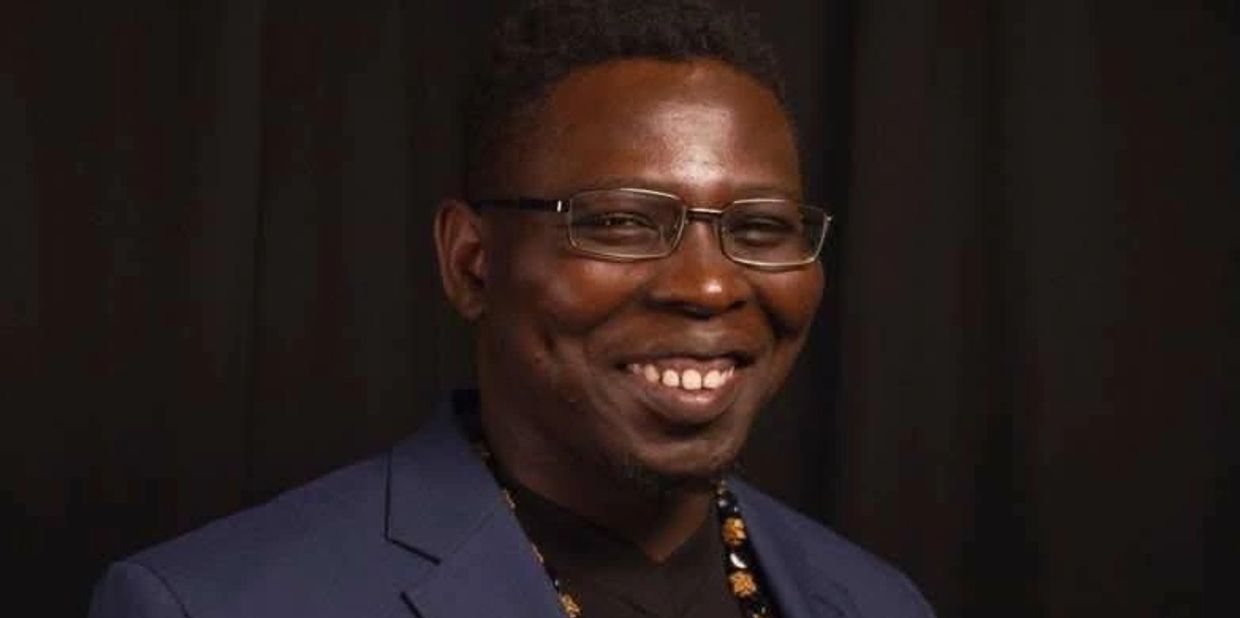 Smiling man wearing glasses and a blue blazer against a dark background.