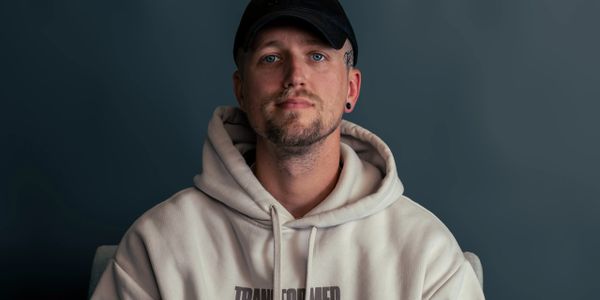 Man in beige hoodie and black cap against a dark background.