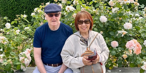 An elderly couple sitting on a bench surrounded by blooming flowers.