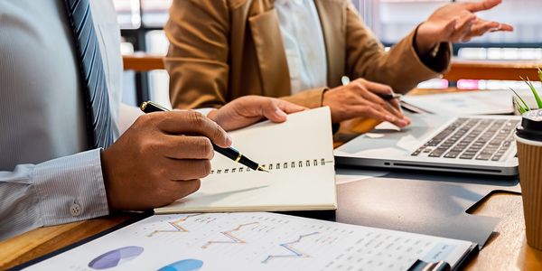 Two people discussing with notebook and laptop on the desk.