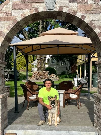 Man in neon shirt kneels with a German Shepherd under a stone arch in a backyard.