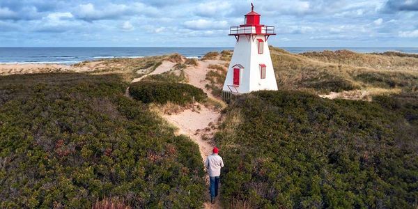 Person walking on a path towards a small red and white lighthouse near the beach.