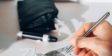 Hand using a pen to fill out a form with a blood glucose meter nearby.
