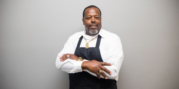 Confident chef in white shirt and black apron posing with crossed arms.