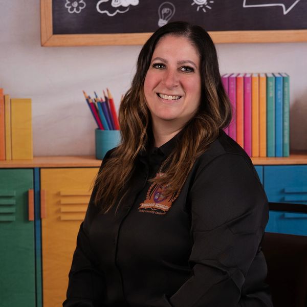 Smiling woman in black shirt poses in colorful classroom setting with books and pencils.