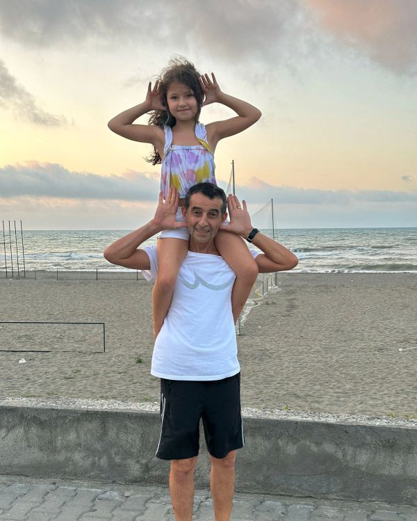 A man with a girl on his shoulders making playful hand gestures by the beach at sunset.