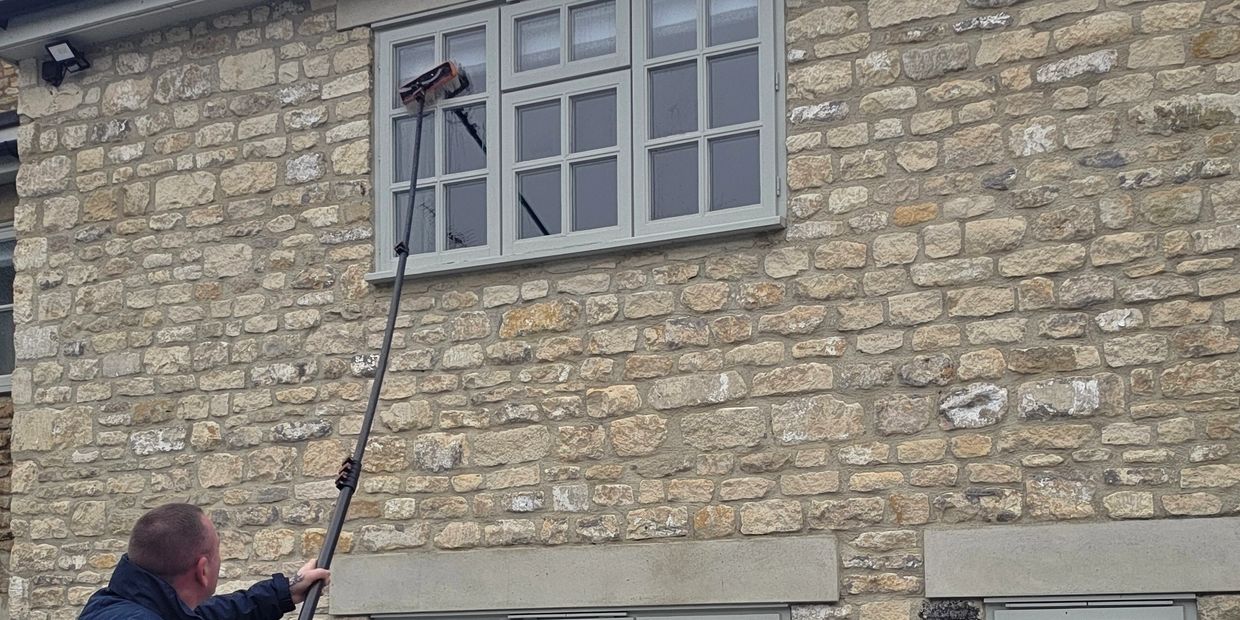 A man cleaning upper windows of a stone house with a long pole.