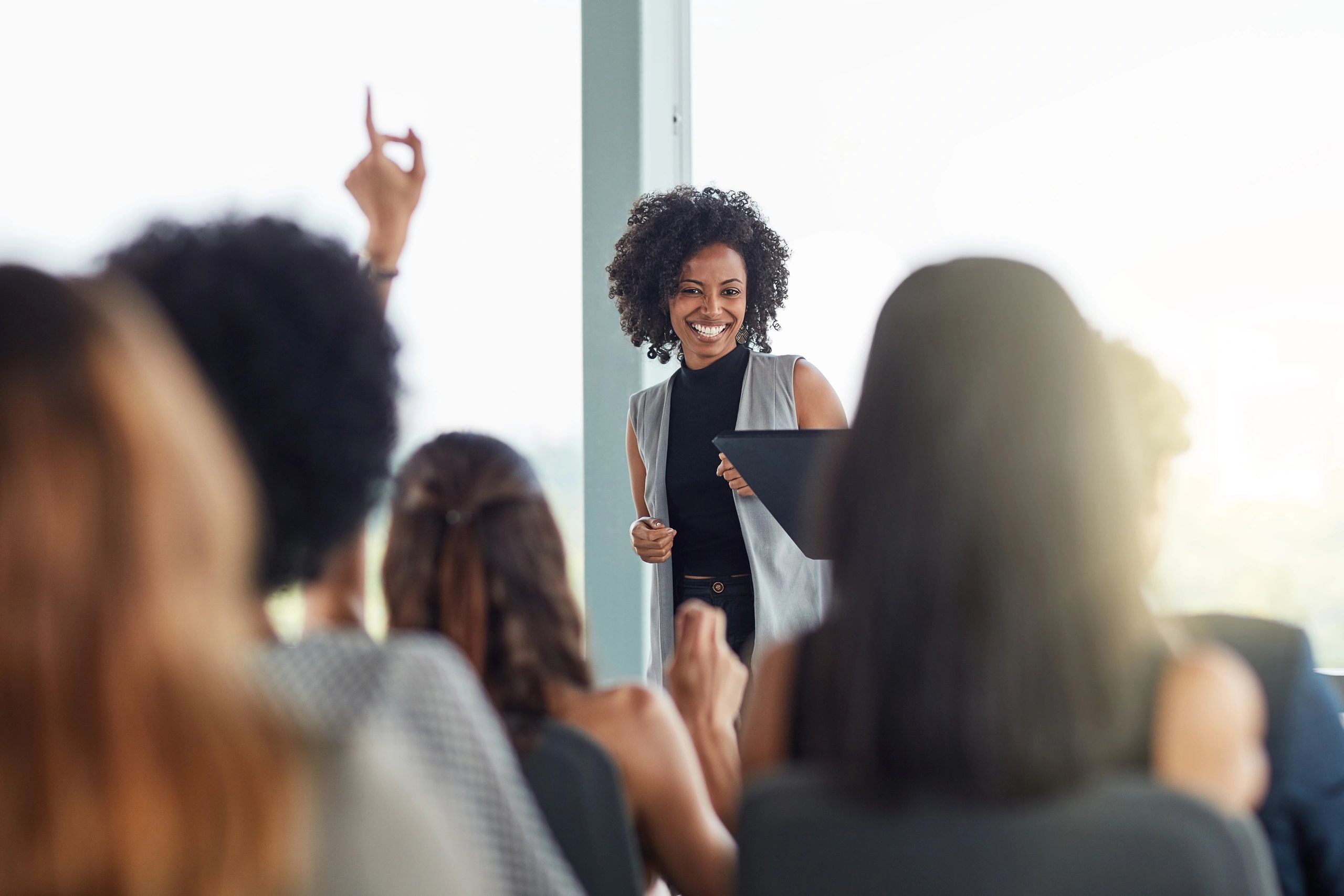 A woman smiles while presenting to an audience with a raised hand.