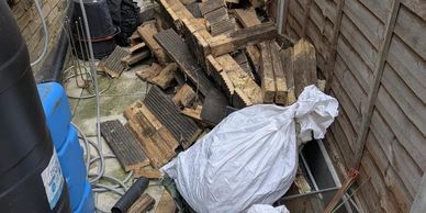 Pile of chopped wood and white bags in a narrow outdoor space beside a brick wall and wooden fence.