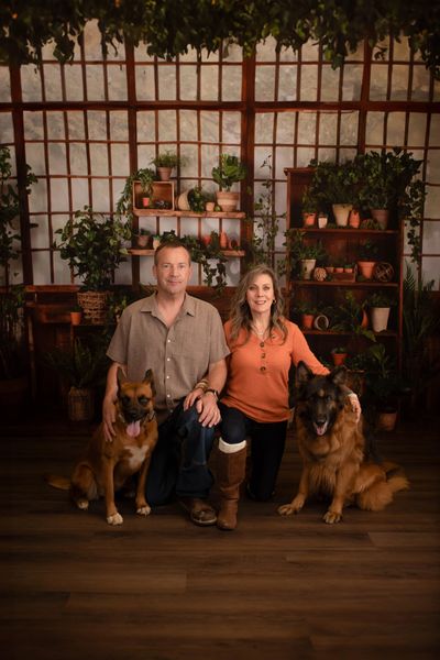 A couple kneeling with two dogs in a cozy plant-filled room.