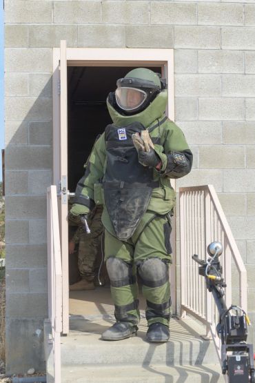 Person in a bomb disposal suit standing at a doorway, holding a device.