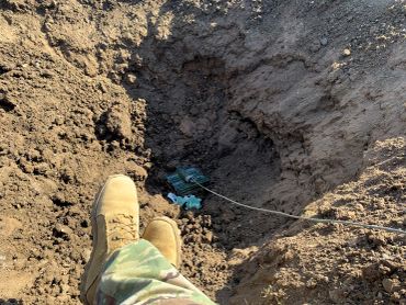 Soldier's boots near a deep hole with a blue object inside in dirt.