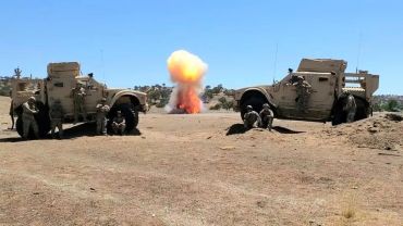 Military vehicles firing artillery in a desert landscape with explosion.