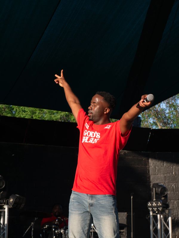 Man in red "God's Plan" shirt with arms raised, holding a microphone.